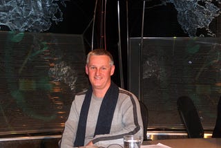 A photo of Ron (a man with salt-and-pepper hair wearing a grey sweater) behind the desk on the set of 22 Minutes in Halifax.