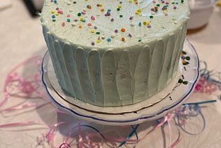 A mint green frosted cake with rainbow sprinkles on a cake stand with ribbons underneath