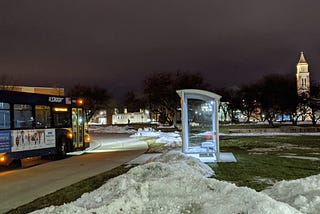 A blue and yellow SMART bus approaches a shelter at night with Oakland University’s clock tower in the background