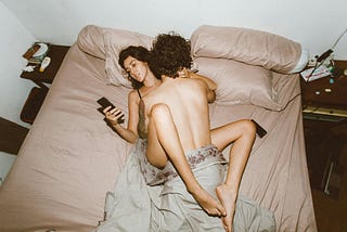 A couple is intimate with one another while the female uses a smartphone.