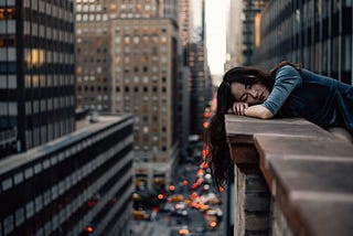 A Woman resting her head on a tall buildings ledge above a busy street