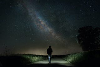 man walking on concrete road between green grass under starry sky during nighttime