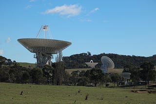 Deep Space Network antennas at Canberra Deep Space Communications Complex in Canberra Australia. The antenna in the foreground is pointed straight up at the sky. Two background antennas are pointed in different directions.
