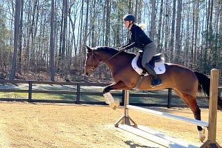 A white woman on a brown horse jumping over a white and brown jump in a sandy arena.