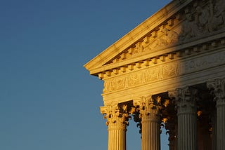 The Supreme Court of the United States building. It is lit by the setting sun, with a shadow cast on one side. The sky in the background is clear and empty.
