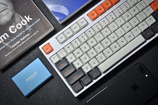 A white mechanical keyboard arranged side-by-side with a portable SSD, books and an iPhone.