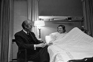 Mrs. Betty Ford sitting up in bed with President Gerald Ford seated to her right. His left hand is holding her right hand.