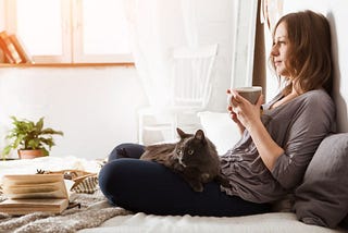 Young smiling woman sitting on a bed, sipping tea with a cat on her lap and a book by her feet | Neurodivergent Life