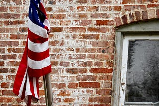 An American flag is tangled around a metal pole in front of a brick wall marked by white paint and a window with peeling frame.