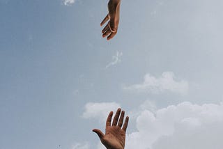 ao fundo o céu, em primeiro plano, duas mãos estendidas uma para a outra de forma vertical.