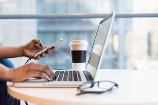 Desk with a laptop, coffee cup, glasses, and a hand holding an iphone.