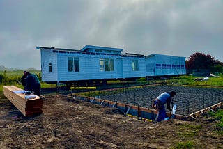 Foundation under construction in foreground and two parts of a manufactured housing unit in background ready for installation. One man working on foundation and two men looking at plans, none with faces visible. Cloudy sky. Trees in distance. Location is Bolinas mesa.