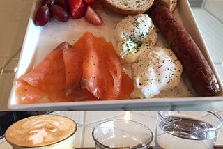 Smoked salmon, eggs, sausage and toast with two glasses of coffee and a glass of water in the foreground.