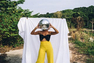 Woman in yellow pants with a disco ball in front of her face as though it’s her head.