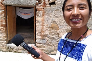 A woman journalist stands with a microphone outside a rural stone house while on a reporting trip in Oaxaca, Mexico.