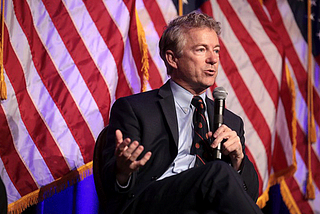 Senator Rand Paul sitting down with a microphone in hand with an American flag behind him.