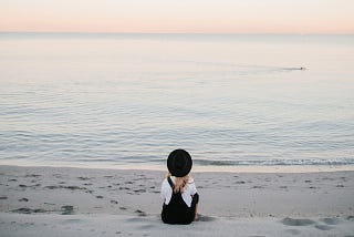 Mim gazes into the ocean. Her back is to the camera, which faces the still water at dawn. There is a person swimming far in the background. The lighting is pastely and gentle.