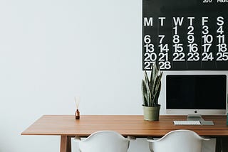 Desk with an iMac to the right of the image with a large black-and-white calendar hanging overhead. Two chairs against the desk. On the desk next to the iMac is a potted plant.