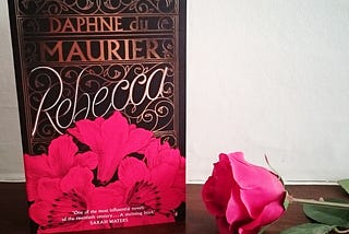 image shows a book — Rebecca by Daphne du Maurier, standing on its end beside a red rose.