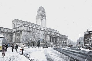 The Parkinson Building including the large clock tower, viewed from Woodhouse Lane. The paths and buildings are lightly covered in a recent snow shower. People are walking up the street.