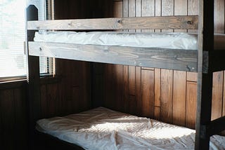 A photo of a wooden bunk bed with bare mattresses and a snipped of window.