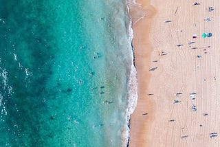 overhead drone shot of beach, turquoise sea at left, sand at right, waves breaking on shore, people dotted along the sand