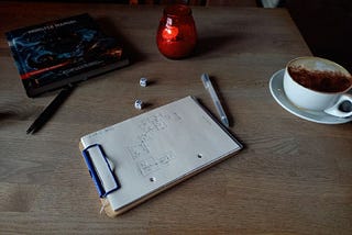 photograph of wooden table with red candle, clipboard with pen drawing, two dice, one pen, one coffee (cappuccino, chocolated on the closest side) and one copy of the Monster Manual