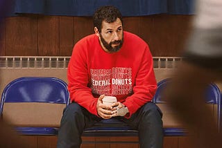 Adam Sandler sentado no banco de reservar de um ginásio de basquete, durante um treino, como protagonista do filme Arremessando Alto, da Netflix.