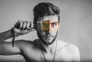 In a black-and-white photograph, an individual wipes a paintbrush across his own face, revealing real-world color.