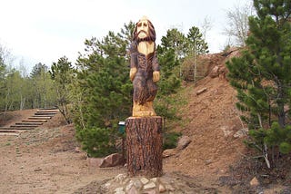 A wooden sculpture of bigfoot sits between a two trees on a hill