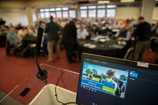 A laptop screen on top of the lectern at the firearms instructors conference behind which there are the attendees sitting at tables (this part of the picutre is blurred). The laptop is showing a slide of a presentation with the text ‘Firearms Instructors Conference — Guardians of the Standard’, the slide has the picture of a group of firearms instructors in the background, set outside in a teaching setting.