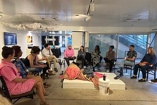 A group of people gather in chairs in a roundtable format in a white room with floor to ceiling windows. They wear bright pinks and dark colors. In the center of the table and overhead are tapestry-based sculptures.