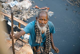 An old man next to a polluted river looks at the camera while holding his cane.