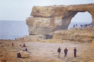 Tiny figures of people stand before a large sandy stone arch, the sea and sky behind them