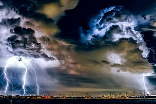 Photo of a storm darkened sky with mixtures of Dark blue, brown, black, and yellow coloring the stormy sky, while bolts of lightning erupt from storm clouds to touch the darkened earth of the city.