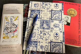 A Tarot card deck, a Tarot workbook, a blue pen on top of a planner, and a copy of Alexander Chee’s essay collection, How to Write an Autobiographical Novel. The card deck is named “Modern Hue” and features watercolor illustrations.