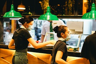 Picture of servers wearing masks picking up food at a counter with green swinging lights overhead, Picture