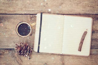 An open notebook with blank pages next to pine cones and a cup of coffee over a wooden table. Glitter overlies, inviting to annual reflection during Christmas time.