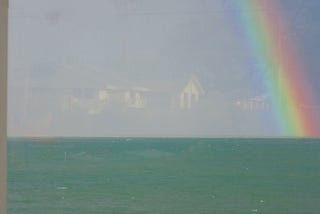 Image of a rainbow a green ocean with a reflection of houses in the sky