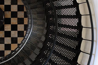 View from above of a black spiral staircase against a white wall, and a black and white checked tile floor