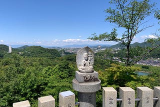 A layered view from a temple at the top of Mt. Tsugao. There is a bright blue sky with picturesque clouds and lush green mountains surrounding a small town in the background. In the foreground is the lookout spot from which the photo is taken, with square stone pillars and a statue of Bodhisattva sitting in front of a leaf-shaped wall with a snake carved into the block beneath him.