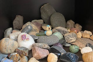 a collection of rocks of different shapes, colors, and sizes, with a tiny bluebird figurine and a trinket of a dragon hatching from an egg.