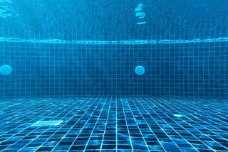 dark tiled swimming pool viewed from the pool floor underwater, with the reflection of the tiles visible in the rippling surface of the water.