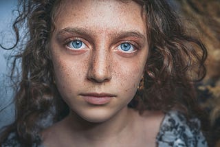 A girl with curly hair and piercing blue eyes looks into the camera with a neutral expression.