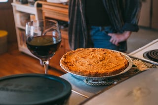 Person standing by a pumpkin pie and glass of red wine.