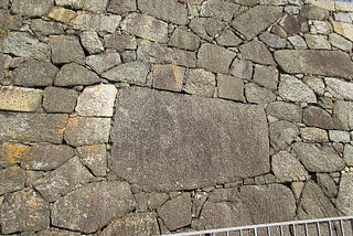 Edo castle wall in Tokyo Japan. There are stones of various shapes are piled up. It is piled up neatly and tightly without any gaps. 
 
 The large stone in the center is several meters. and the others are made of small stones of about 50 cm. 
 
 The image represents is the following things. The accumulation of people’s lives creates a society. Create a future society with strong harmony. A slightly blue sky can be seen in the upper right. It shows hope in the future society. society.