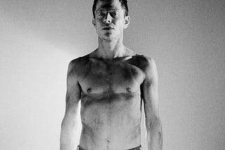 A petite, slightly muscular shirtless white man stares intensely at the camera. The photo is black and white.