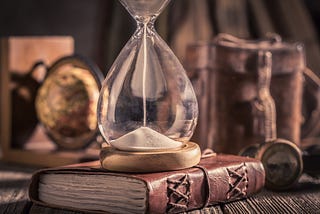 A centuries old hourglass sits atop an old leather-bound book on a desk.