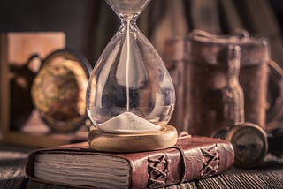 A centuries old hourglass sits atop an old leather-bound book on a desk.