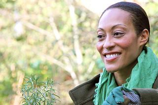 Woman in green scarf smiles off the camera with a background of trees.
